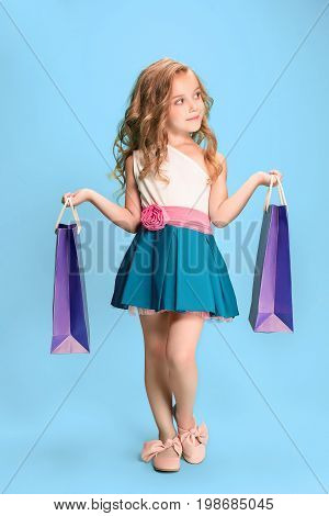The cute little caucasian brunette girl in hat holding shopping bags on blue studio background
