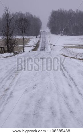 Vertical of a long snow covered country road framed by bare trees and leading a distant tree line in Akwesasne, New York on an overcast blustery day in February.