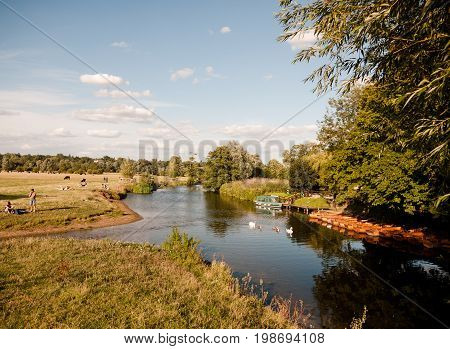 A Summer Scene In Dedham With The River Stour, Swans, Boats, And People Relaxing And Walking