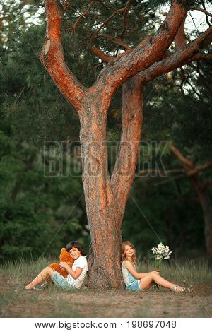 Little girl and boy sit in forest under pine tree. Boy is holding teddy bear in his hands and girl is flower. First love.
