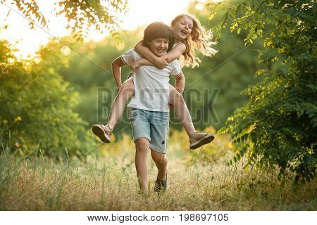 Little boy carries girl on his back in forest. They laugh fun. First love.