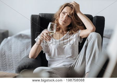 Favourite wine. Cheerless pensive nice woman holding a glass of wine and thinking about her problems while sitting in the armchair