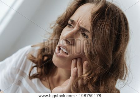 Pain in the tooth. Moody upset unhappy woman holding her cheek and thinking about visiting a dentist while having a tooth inflammation