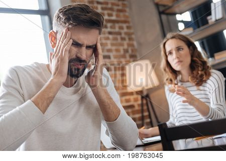 Stop shouting. Unhappy nice handsome man holding his temples and rubbing them while having a quarrel with his girlfriend