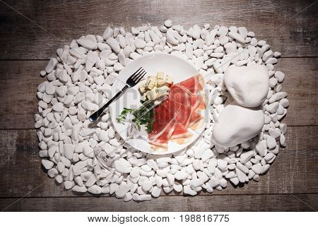 A view from above on a plate with sliced balyk, cheese pieces and fresh leaves on a light wooden table background. A plate with restaurant ingredients and a fork on white rocks.