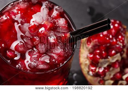 A top view of a picture of a benefit pomegranate juice with cubes of ice on a gray blurred background. The freshly garnet juice and red garnet berries on the desk. The glass of cocktail with straw.