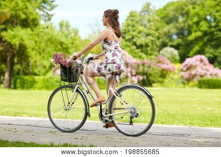 people, leisure and lifestyle - happy young hipster woman wearing summer dress riding fixie bicycle with wild flowers in basket at park