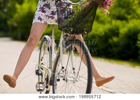 people, leisure and lifestyle - happy young hipster woman wearing summer dress riding fixie bicycle with wild flowers in basket at park