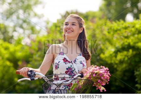 people, leisure and lifestyle - happy young hipster woman wearing summer dress riding fixie bicycle with wild flowers in basket at park