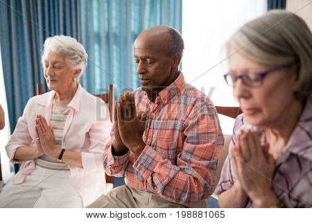 Senior man amidst women praying while sitting on chairs at retirement home