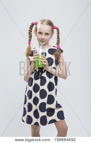 Portrait Of Cute And smiling Caucasian LIttle Girl With Pigtails Posing in Polka Dot Dress with Cup of Greeen Juice. Drinking Through Straw. Vertical Image
