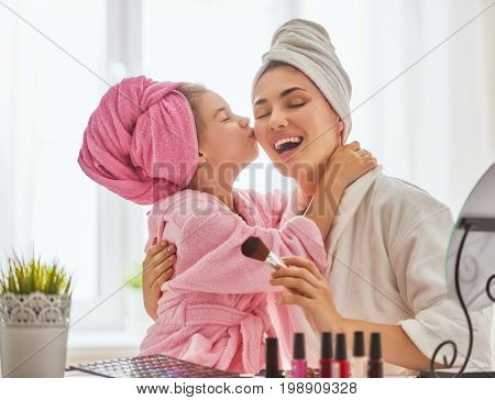 Happy loving family. Mother and daughter are doing make up and having fun sitting at dressing table at home. Mom and child girl are in bathrobes and with towels on their heads.