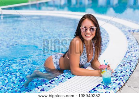 Seductive Girl In Blue Dress Relaxing At Swimming Pool Edge Poolside. Young Sexy Woman With Tropical