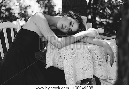 Portrait Of A Young Woman In A Long Evening Dress, She Sits At A Table In The Woods. Black And White
