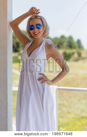 Young beautiful woman in stylish sunglasses stnding in summer house. Outddors portrait of blond lady in white dress.