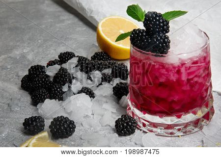 Refreshing cocktail with fresh green mint and sappy blackberries in a pink glass on a grey background, blackberry, slice of lemon and a leaves of mint on a white table-cloth, closeup.
