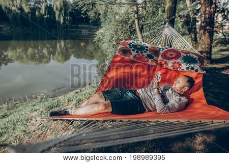 Handsome man relaxing on a hammock on a camping trip stylish hipster man lying on a hammock thinking and looking at phone near a lake relaxation concept