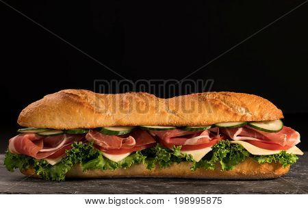 Huge fresh crispy baguette sandwich with meat prosciutto cheese lettuce salad and vegetables. Close up. Black background. Space for text.