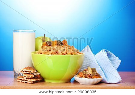 leckere Cornflakes in grüner Schale, Äpfel und Glas Milch auf Holztisch auf blauem Hintergrund