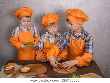 Three Cute Boys In Costumes Cooks Engaged In Cooking Homemade Burgers. Three Brothers Preparing Fami