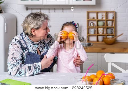 Happy Little Girl And Her Grandmother Have Breakfast Together In A White Kitchen. They Are Having Fu