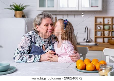 Happy Little Girl And Her Grandmother Have Breakfast Together In A White Kitchen. They Are Having Fu