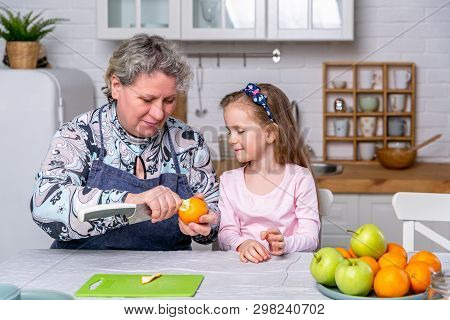 Happy Little Girl And Her Grandmother Have Breakfast Together In A White Kitchen. They Are Having Fu