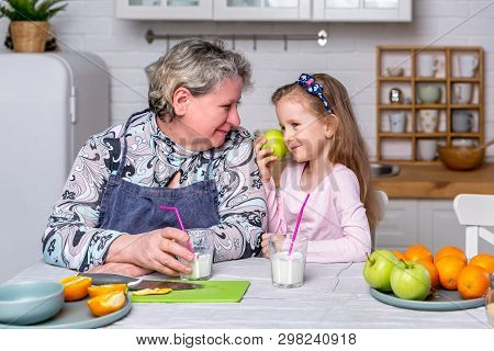 Happy Little Girl And Her Grandmother Have Breakfast Together In A White Kitchen. They Are Having Fu