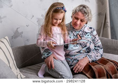 Grandmother And Her Little Granddaughter Are Watching Movies Together And Playing On The Device Whil