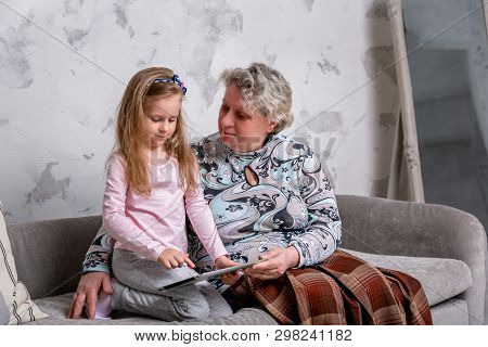 Grandmother And Her Little Granddaughter Are Watching Movies Together And Playing On The Device Whil