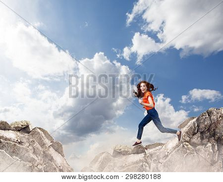 Redhead Woman Jumps Through The Gap Between Hills Over Cliff On Blue Sky Background.