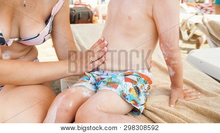 Young Caring Mother With Little Oddler Boy Sitting On Sunbed On Beachand Using Sunscreen Lotion Befo