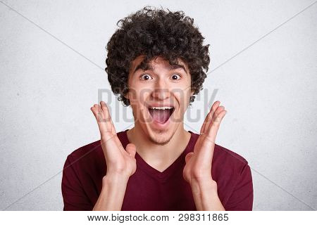 Studio Shot Of Attractive Man With Dark Curly Hair, Keeps Mouth Opened, Screams Something, Stands Wi