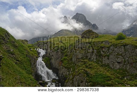 Little Waterfall Among Green Rocks. Massive Shrouded Rock In The Background.