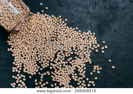 Healthy Food, Dieting And Nutrition Concept. Dry Uncooked Chickpeas Spilled From Glass Jar On Dark B