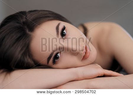 Beautiful Brunette Girl With Brown Eyes With Bare Shoulders Lying On The Bed. Face Close Up. Asian A