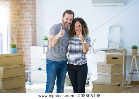 Middle age senior couple moving to a new home with boxes around doing happy thumbs up gesture with hand. Approving expression looking at the camera with showing success.