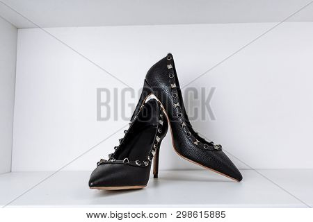 Pair Of Black High-heeled Shoes With Pointed Toes, Decorated With Metal Inserts Against A Shelf In T