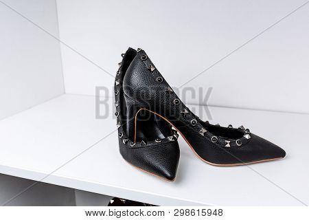 Pair Of Black High-heeled Shoes With Pointed Toes, Decorated With Metal Inserts Against A Shelf In T