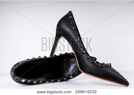 Pair Of Black High-heeled Shoes With Pointed Toes, Decorated With Metal Inserts Against A Shelf In T