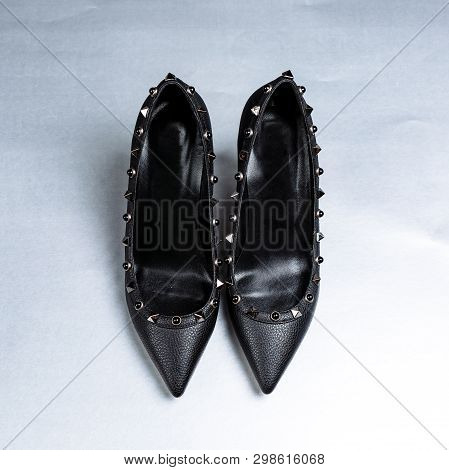 Pair Of Black High-heeled Shoes With Pointed Toes, Decorated With Metal Inserts Against A Shelf In T