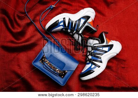 White Sneakers With Black And Blue Inserts On A Thick Figured Sole And A Blue Bag With A Gold Chain