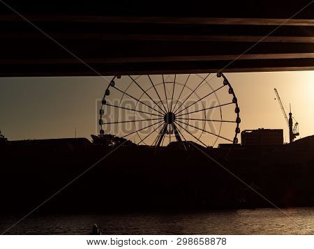View From Underbridge Showing Silhouette Of Riverside Ferris Wheel And Construction Crane.
