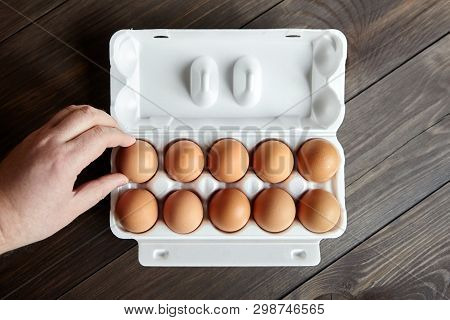 Ten Chicken Eggs In A White Tray On A Brown Wooden Table. Male Hand Takes The Egg From A Box