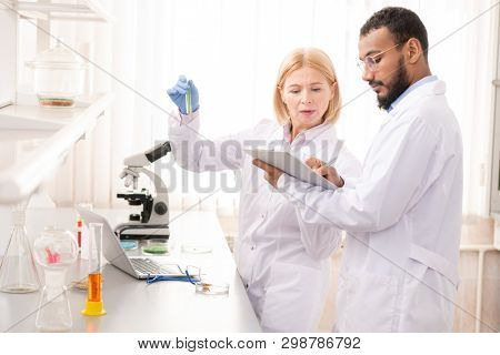 Serious concentrated pharmaceutical specialists studying chemicals while mixing reagents in test tube and checking notes in paper, they working in laboratory