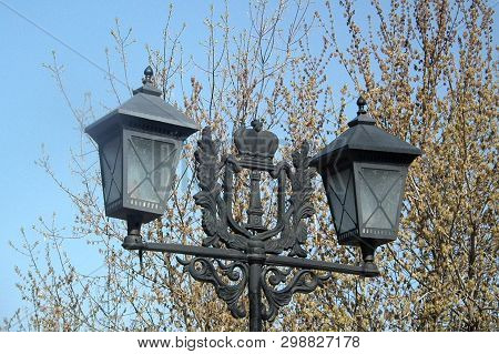 Vintage Vintage Lantern With A Coat Of Arms Against The Blue Sky And Tree Branches.