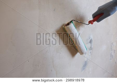 A Male Blue-gloved Workers Hand Applies A Primer To A White Wall Using A Roller. Background Of Wet C