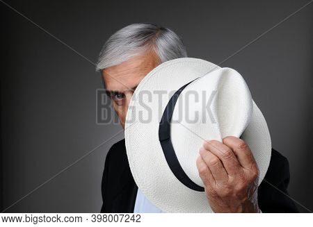 Closeup of a middle aged man wearing a tuxedo and a Panama Hat in his hand as he peeks around the hat. Horizontal over a gray background.