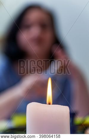 A Blurry Silhouette Of A Brunette Sitting At A Table. In The Foreground Is A Lighted White Candle. T