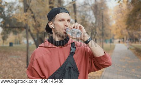 Man Runner Closeup In Red Hoodie And Earbuds Drinking Water In The Park Slow Motion, On Background O